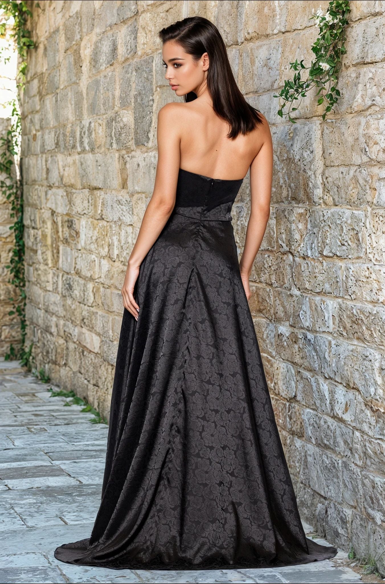 A woman with straight dark hair models the Strapless Black Prom Gown: Blondie Nites 5301BN, standing with her back to the camera against a stone wall covered in green vines.